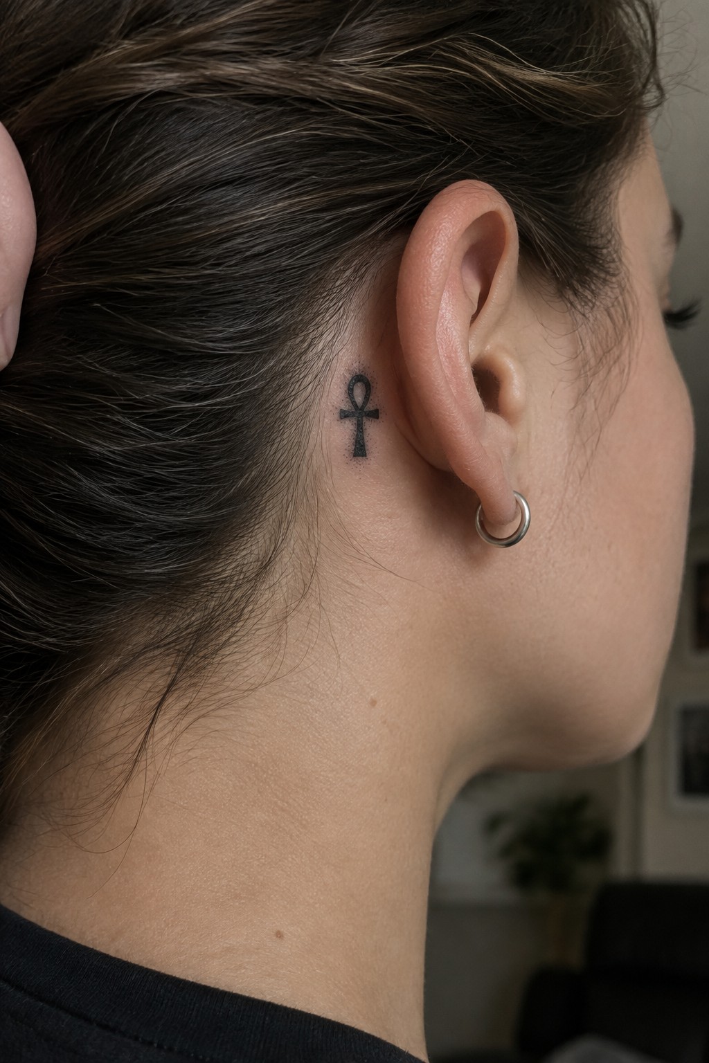 Woman's neck with Ankh tattoo behind ear and silver hoop earring in brown hair.