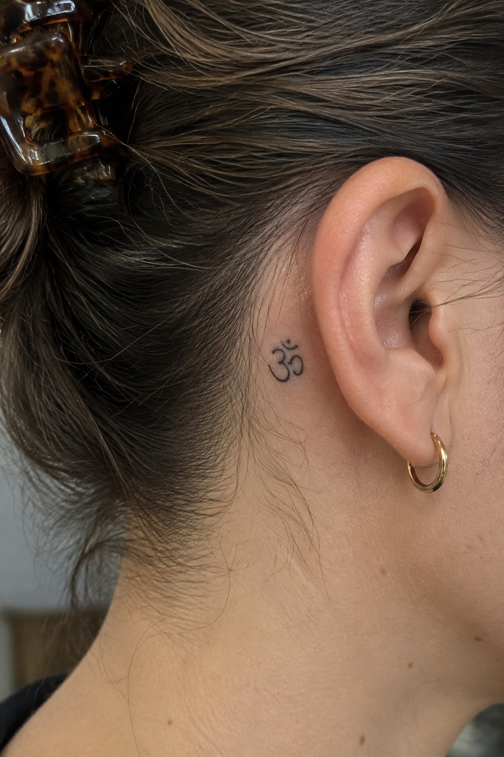 Om tattoo behind ear with gold hoop earring, brown hair in tortoiseshell clip.