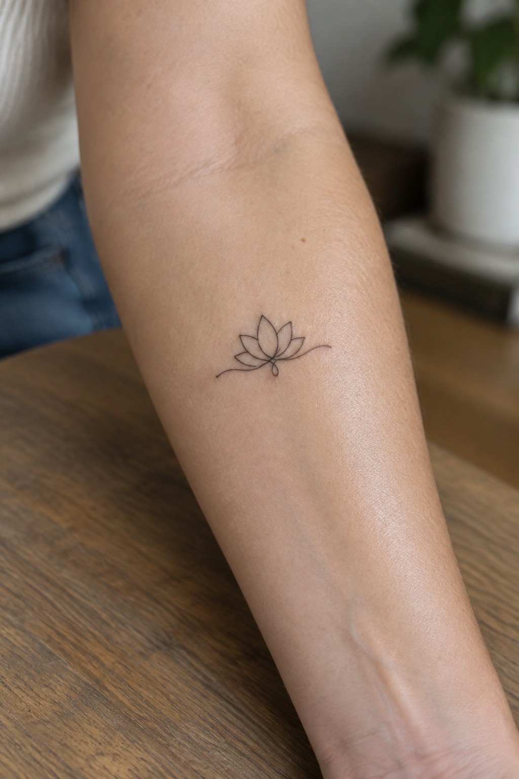 Minimalist lotus flower tattoo on forearm resting on wooden table beside jeans