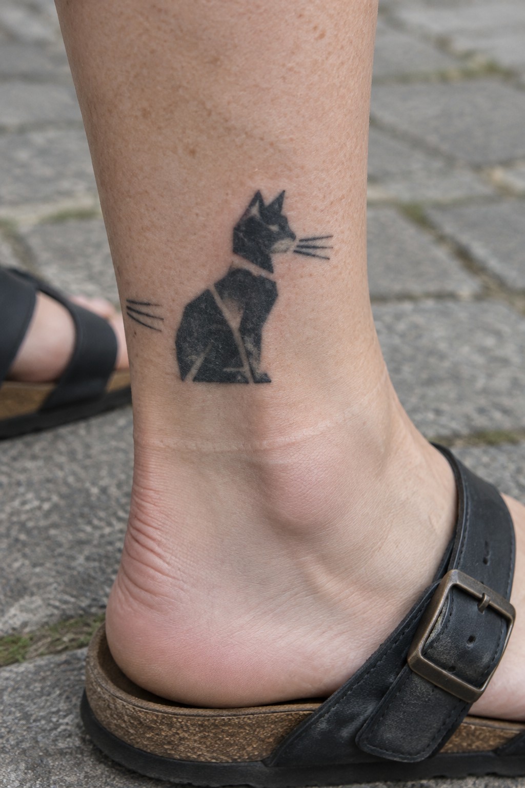Black geometric cat tattoo on ankle above black leather sandal on pavement.