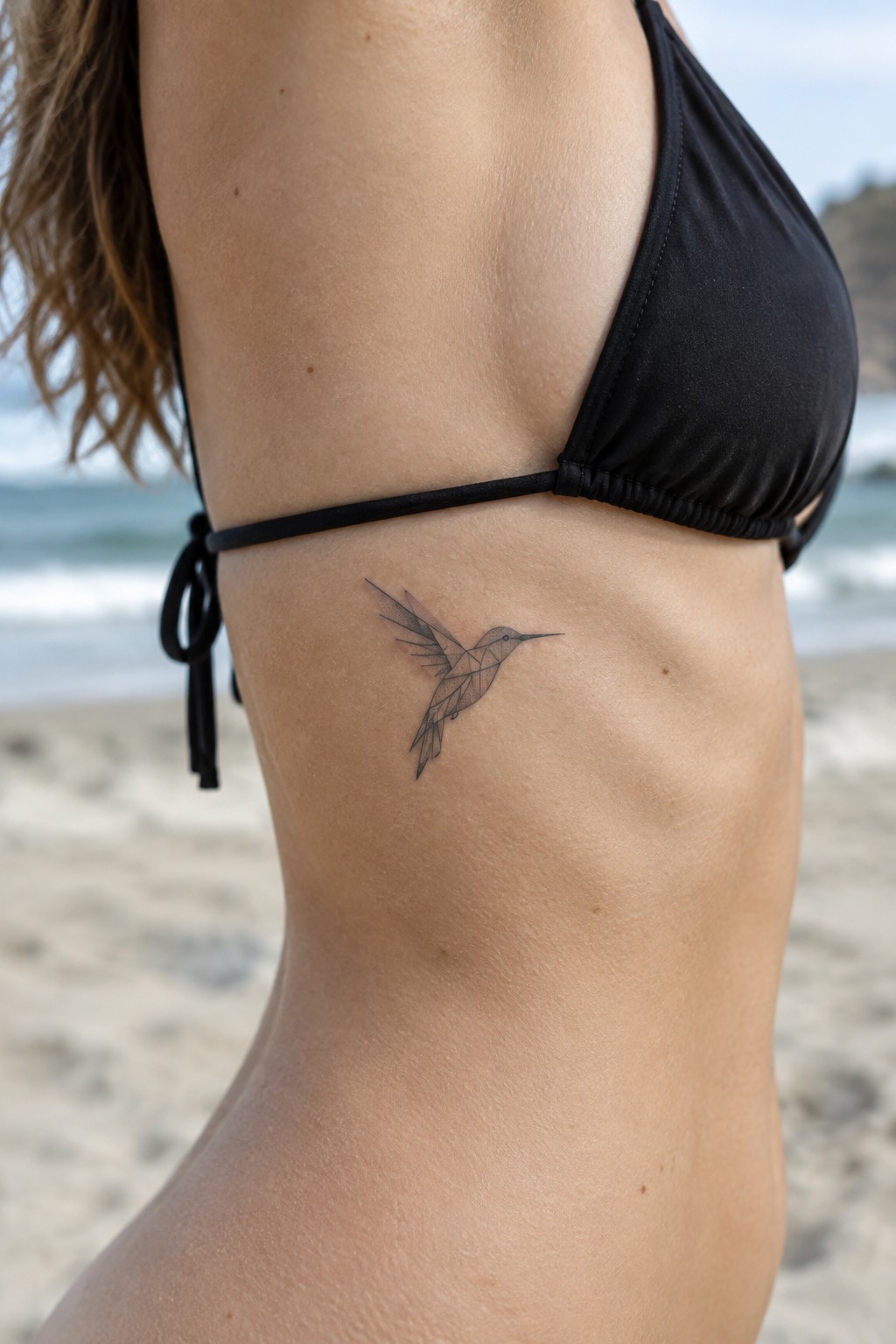 Woman in black bikini with geometric hummingbird tattoo on ribs at beach