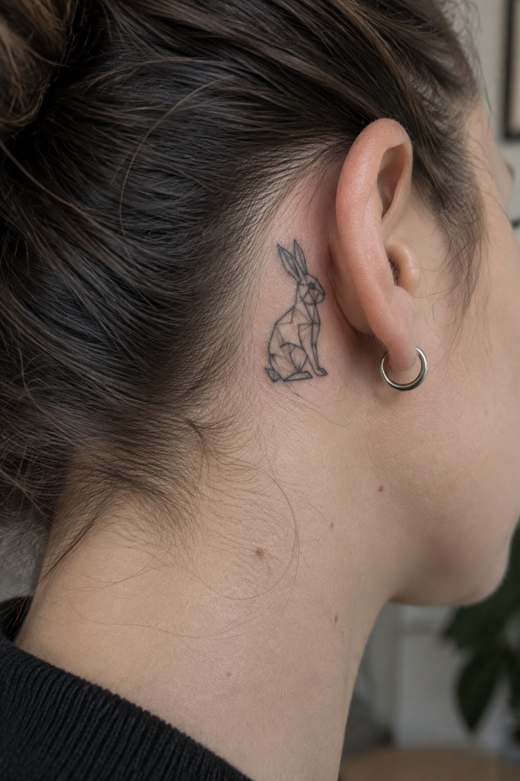 Geometric rabbit tattoo behind ear of dark-haired person with bun and hoop earring.