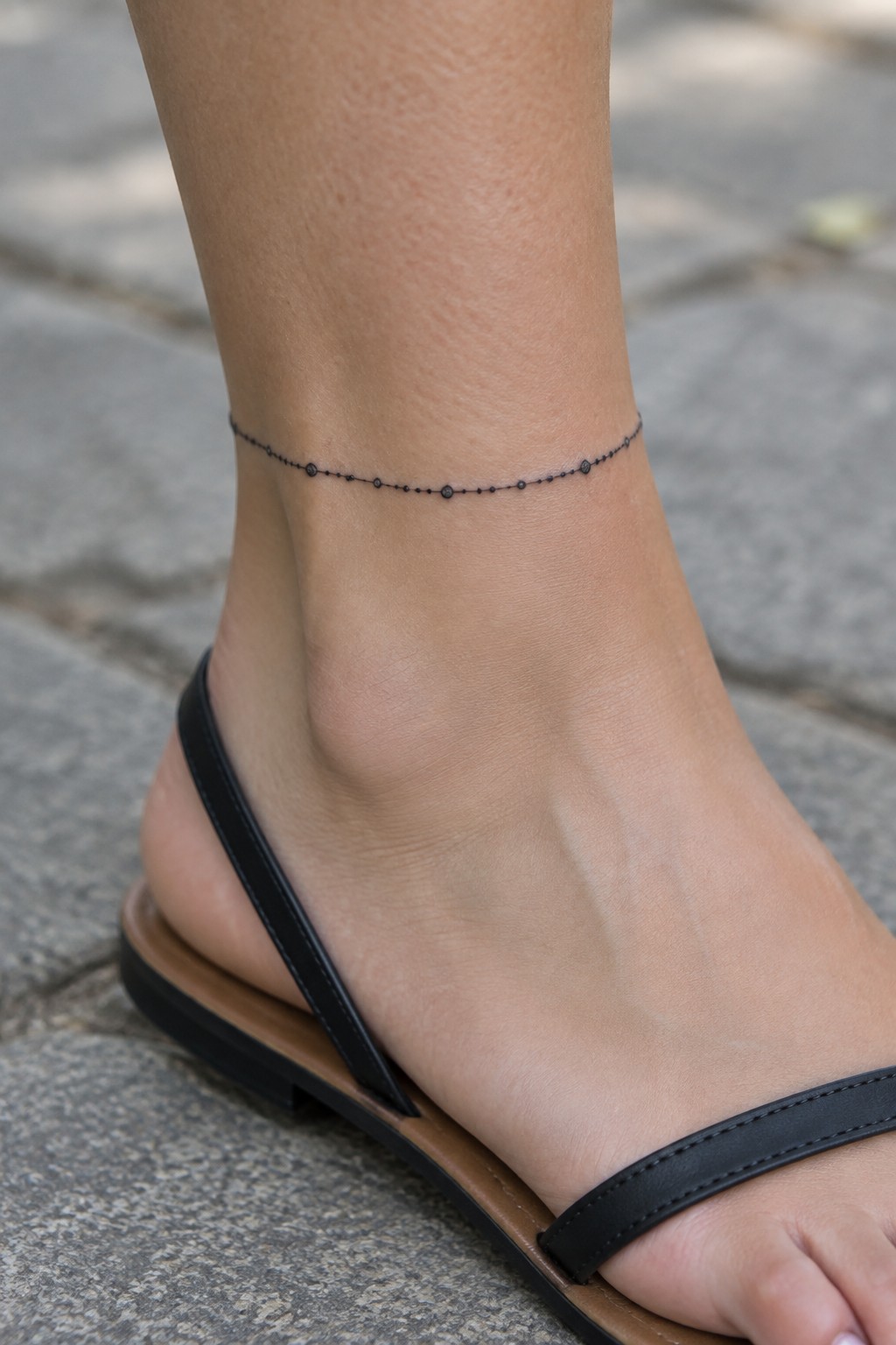 Fine line tattoo of a thin beaded chain wrapped around a woman's ankle above a black sandal on pavement.