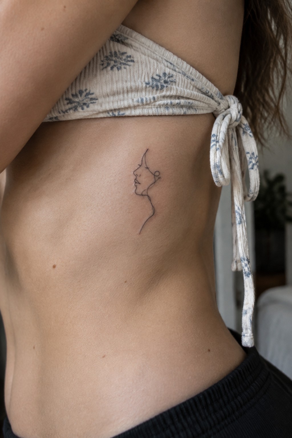 Minimalist line tattoo of woman's profile on ribcage below patterned crop top.