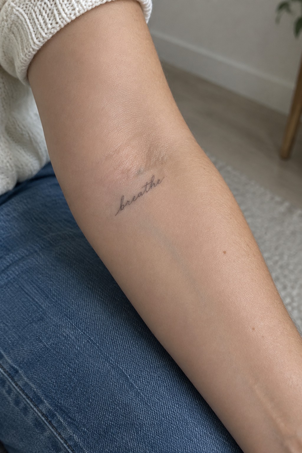 Arm with cursive 'breathe' tattoo, white knit sweater, blue denim jeans visible.