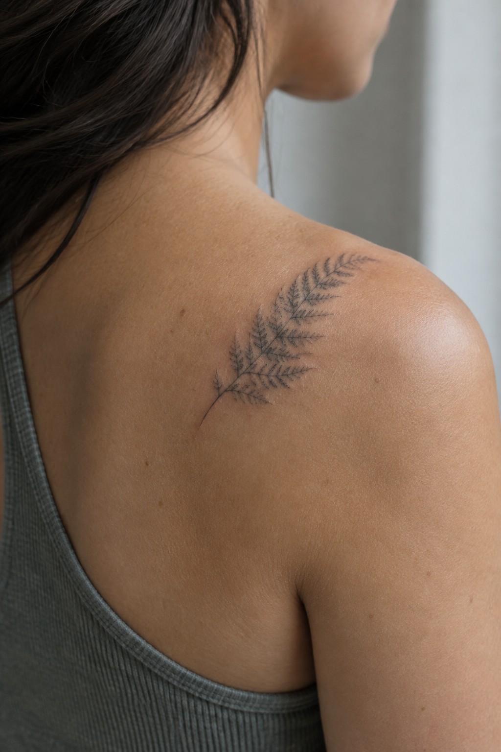 Woman's shoulder with delicate black fern tattoo above gray tank top.