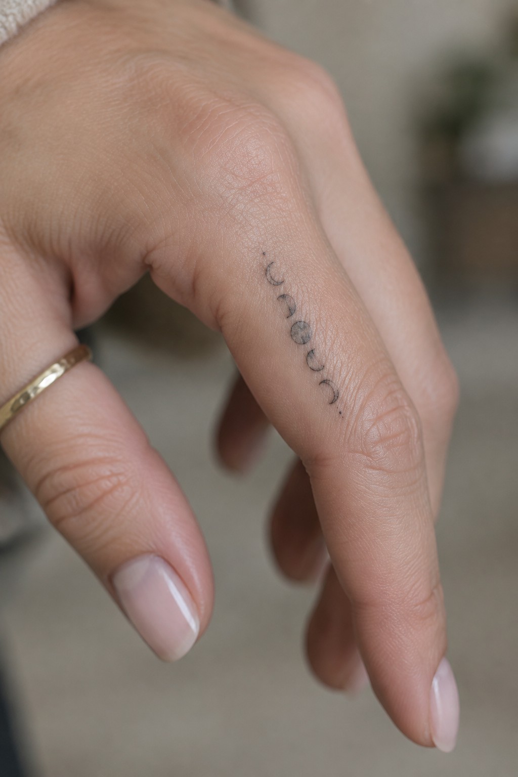 Hand with moon phase tattoo on finger, gold ring, and pale pink manicured nails