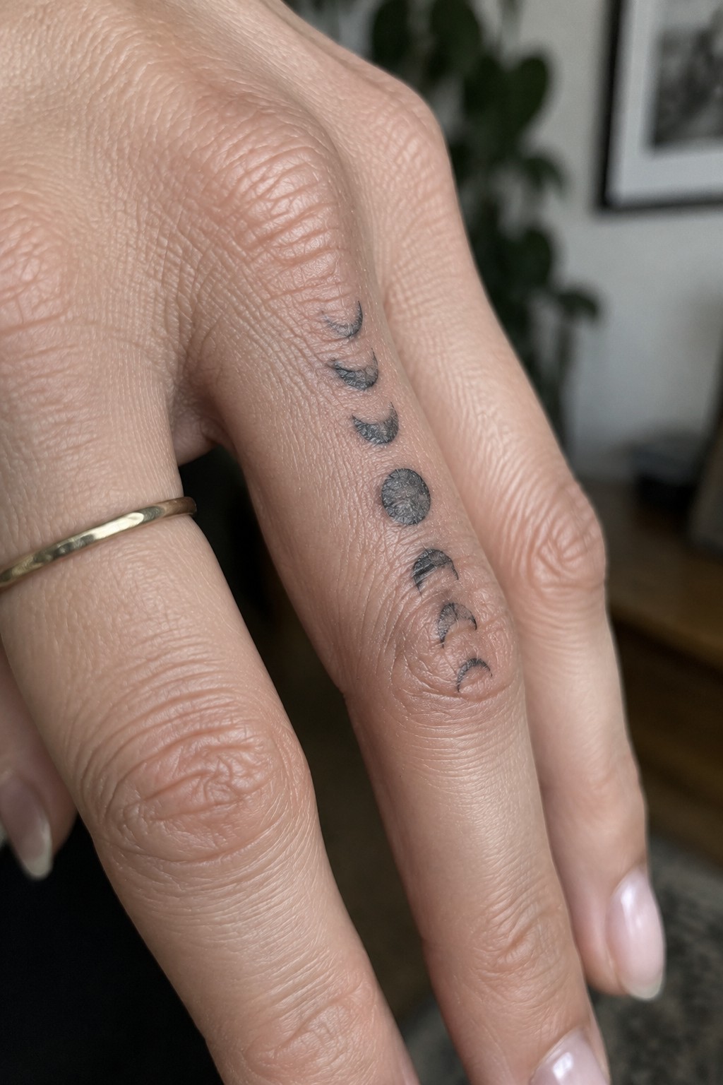 Hand with vertical moon phase tattoo on ring finger and thin gold band.