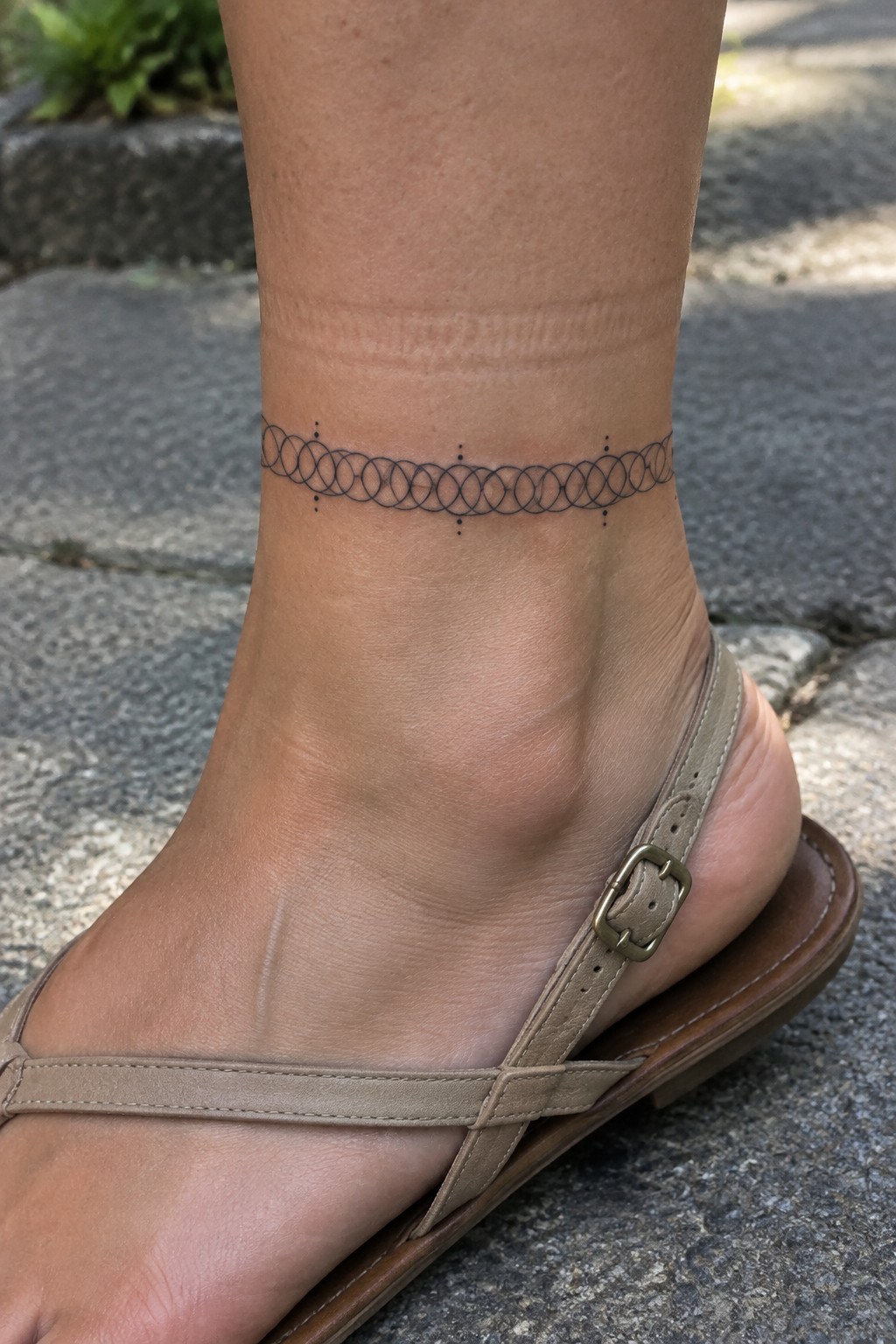 Ankle tattoo of linked circles and dots above beige buckled sandal on pavement
