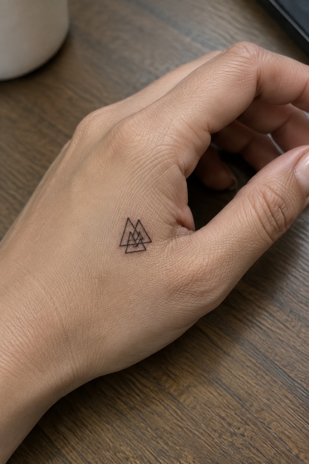 Small black line tattoo of overlapping triangles on the back of the hand near the thumb base