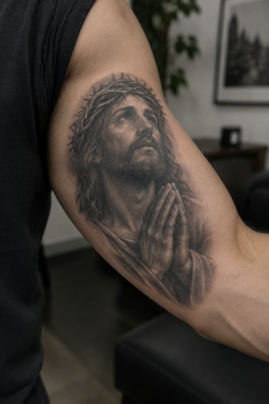 Black and gray realistic tattoo of Jesus wearing a crown of thorns with hands clasped in prayer on a man's upper arm