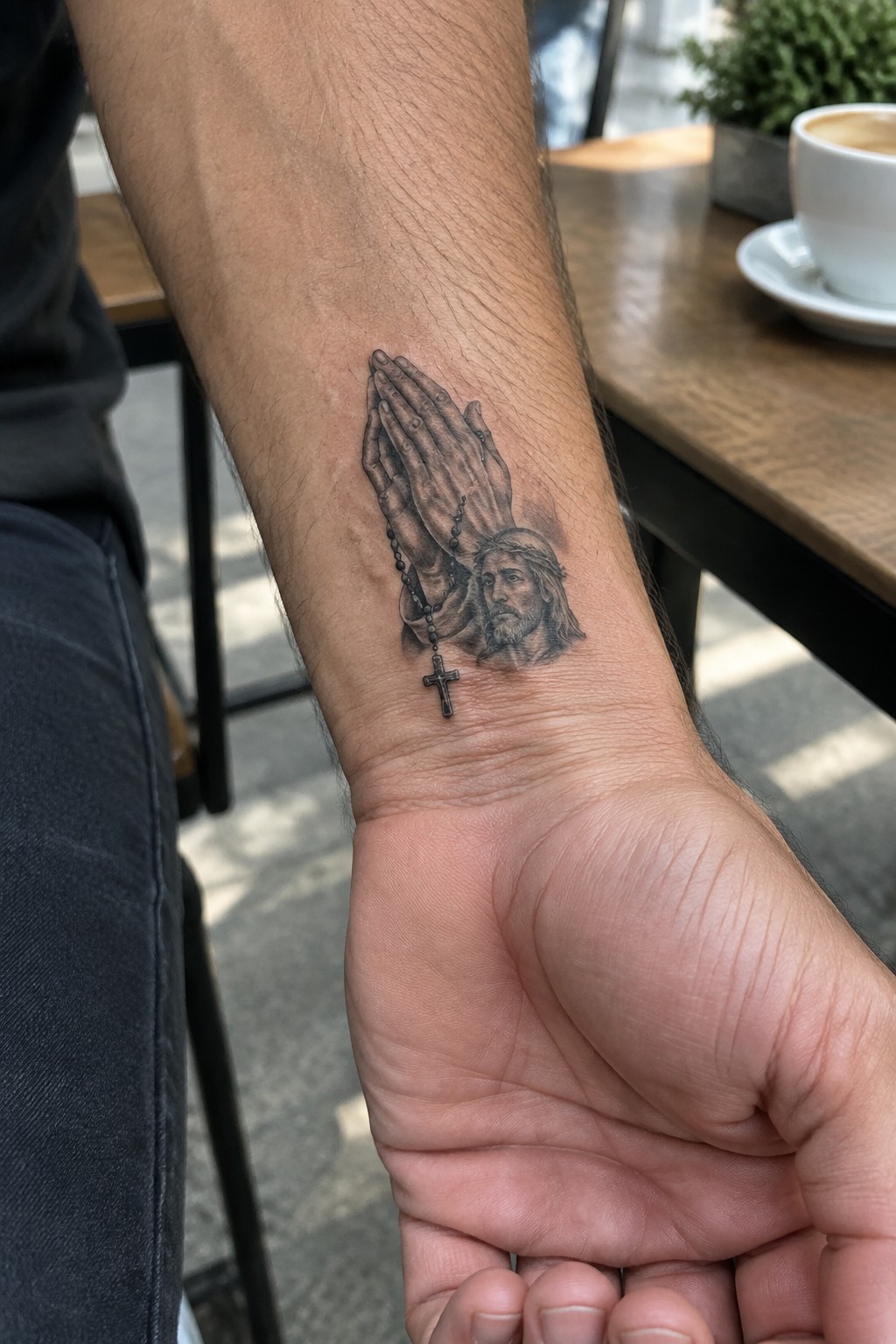 Black and gray realism tattoo of praying hands holding a rosary with a Jesus face on a man's inner forearm near the wrist