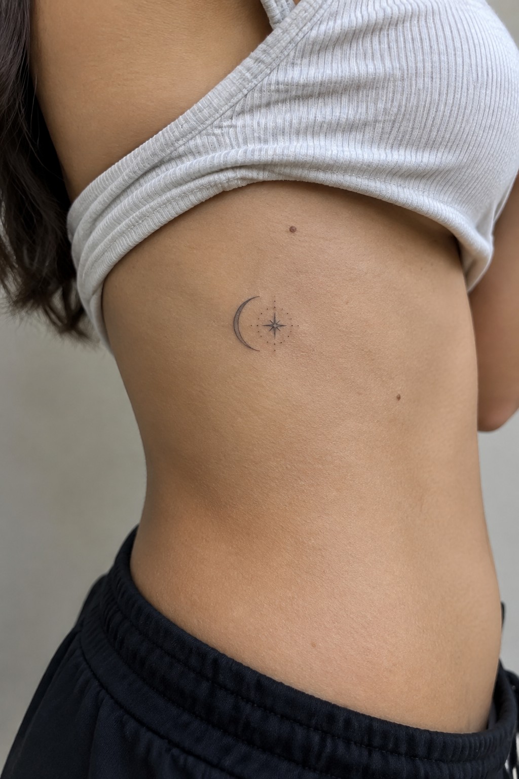 Small crescent moon and star tattoo on torso under white ribbed crop top.