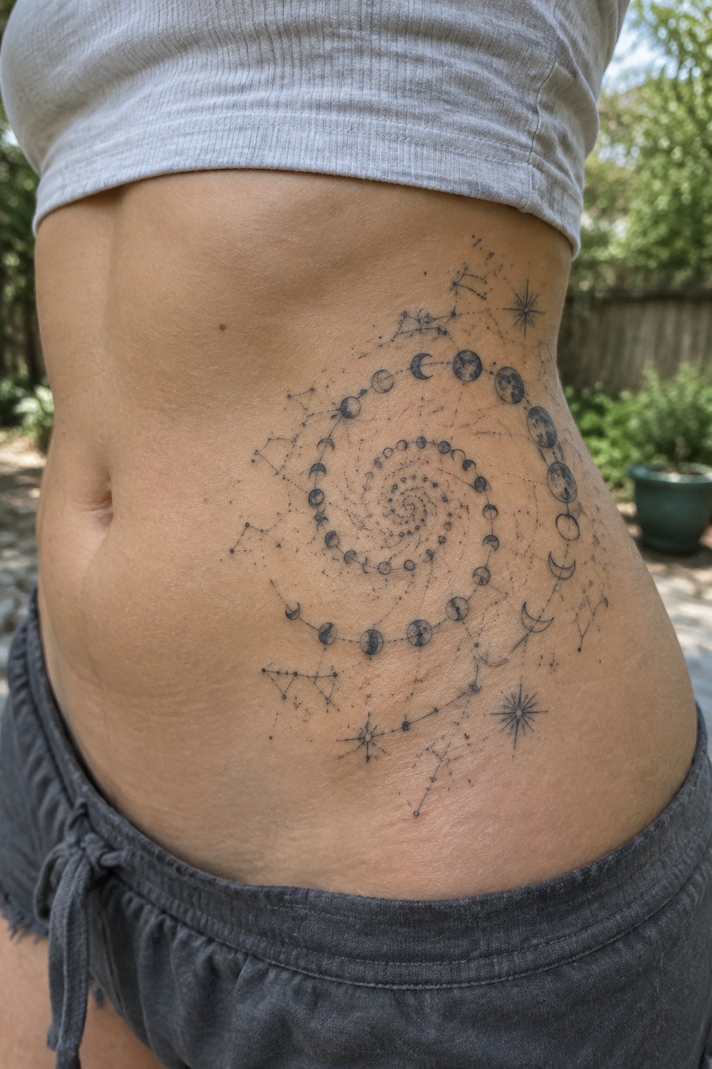 Spiral moon phase tattoo with constellations on midriff in gray crop top and shorts