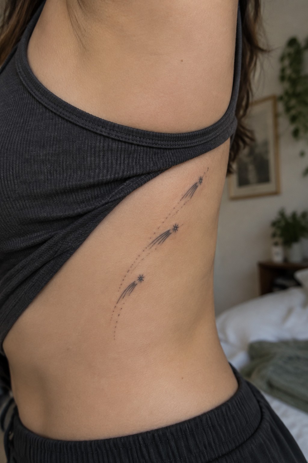 Shooting star tattoos on ribcage under lifted gray ribbed tank top in bedroom