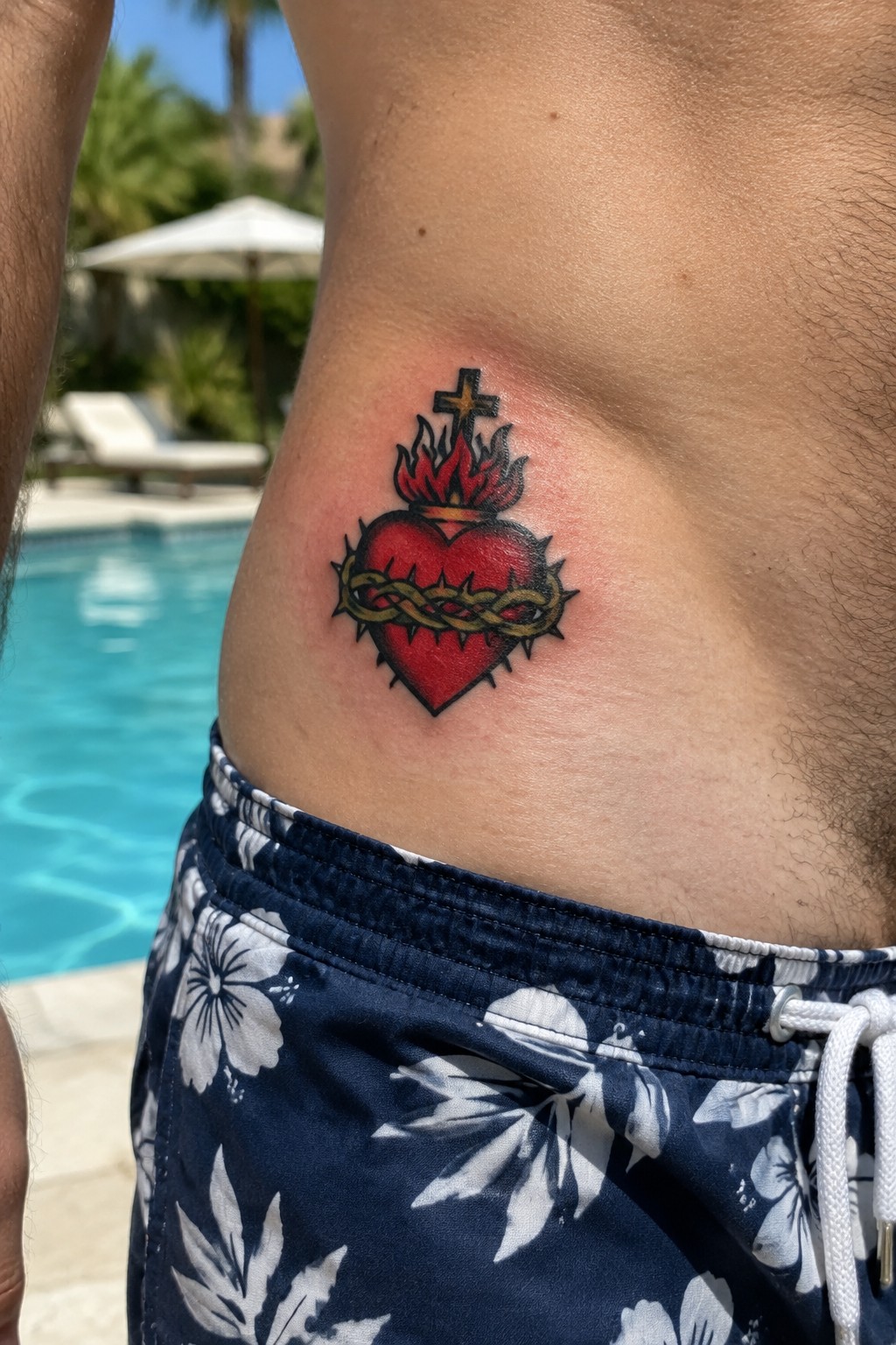 Sacred heart tattoo with red flames, cross, and thorns on side above floral swim trunks