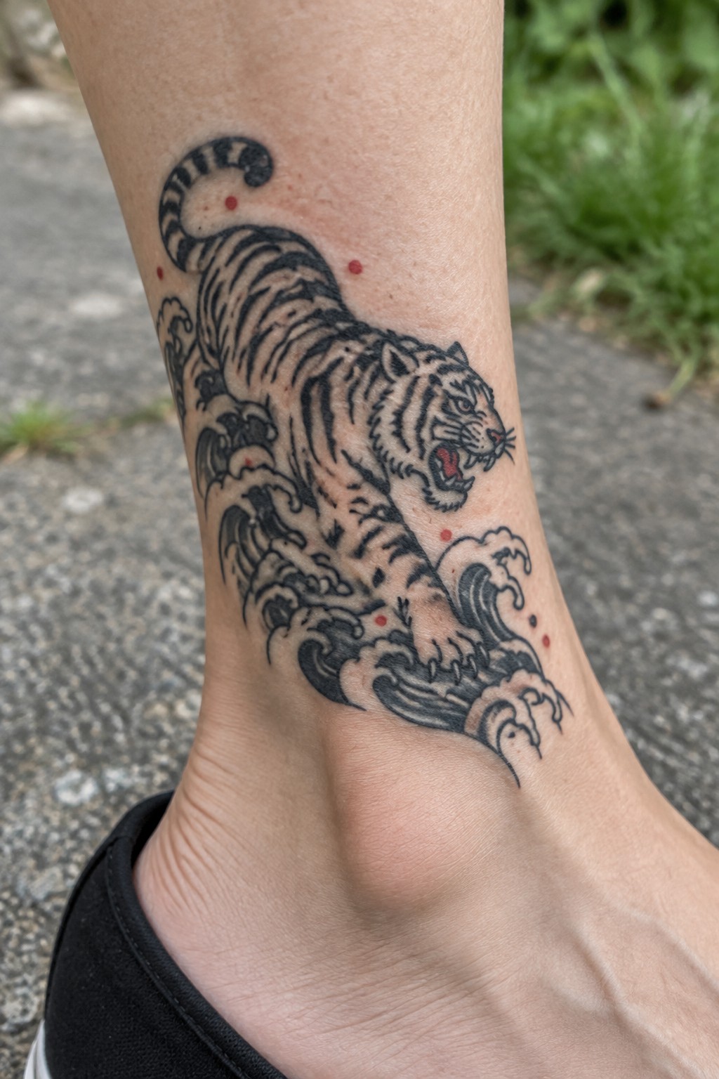 Roaring tiger tattoo with waves and red dots on ankle above black shoe.