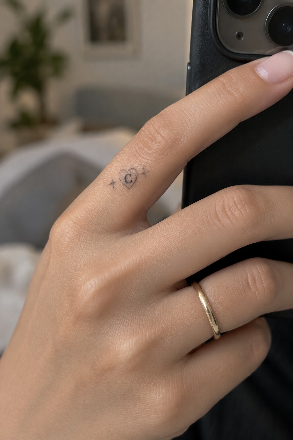 Hand with heart-C tattoo and stars on finger, gold ring, holding black phone