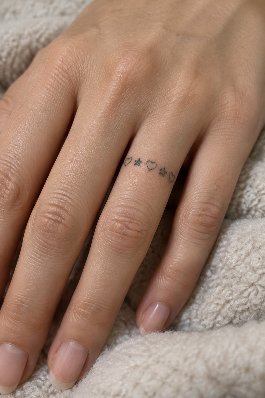 Fine line tattoo of alternating small hearts and stars along the side of a ring finger on a light skin tone hand resting on white fabric