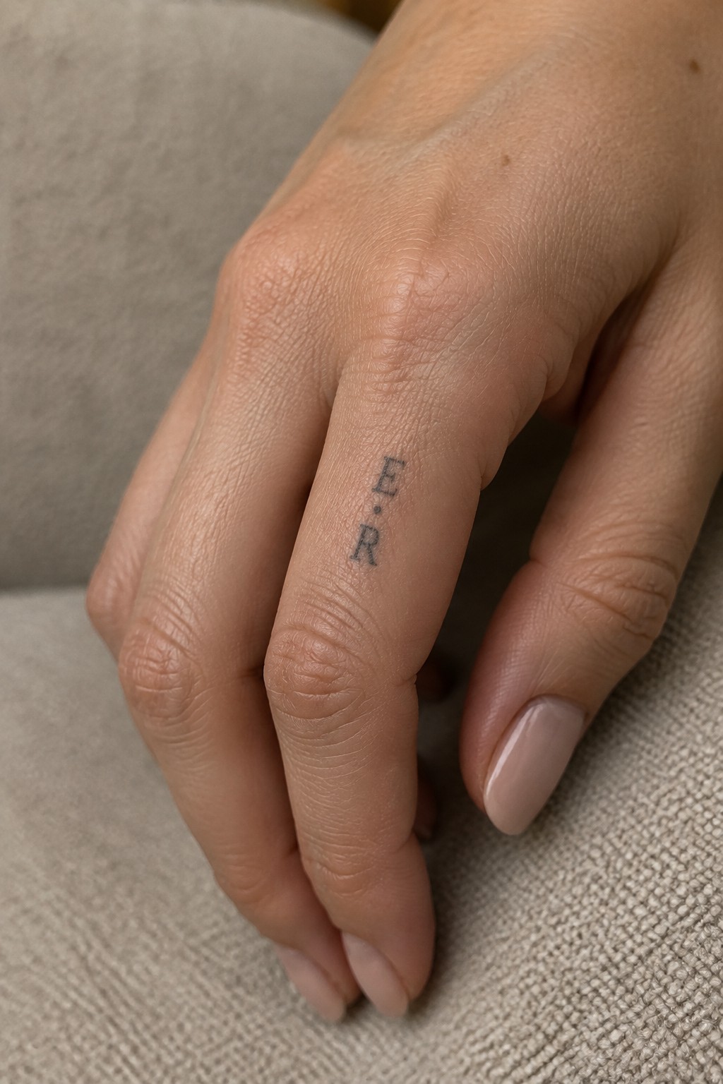 Close up of fine line initials E and R tattooed vertically on the side of a finger with a small dot between them.