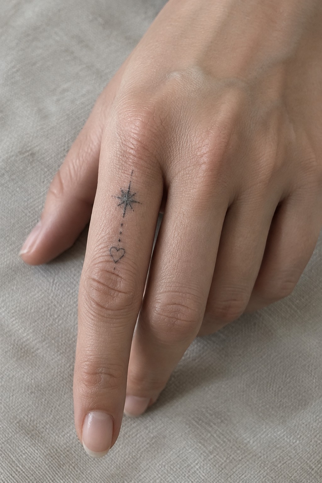 Hand with black tattoo of starburst, dotted line, and heart on finger.