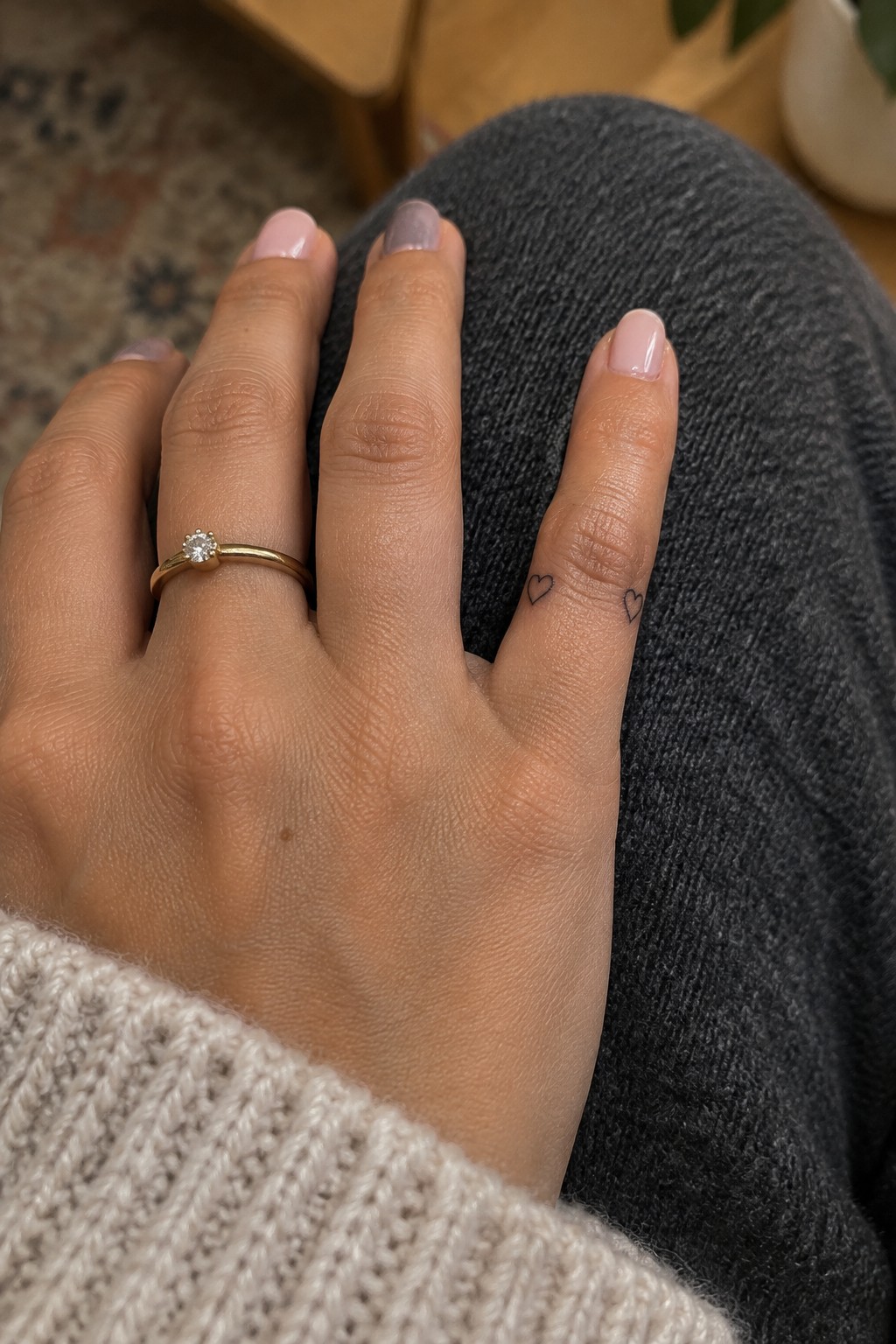 Hand with gold diamond ring and heart tattoos on pinky resting on gray jeans