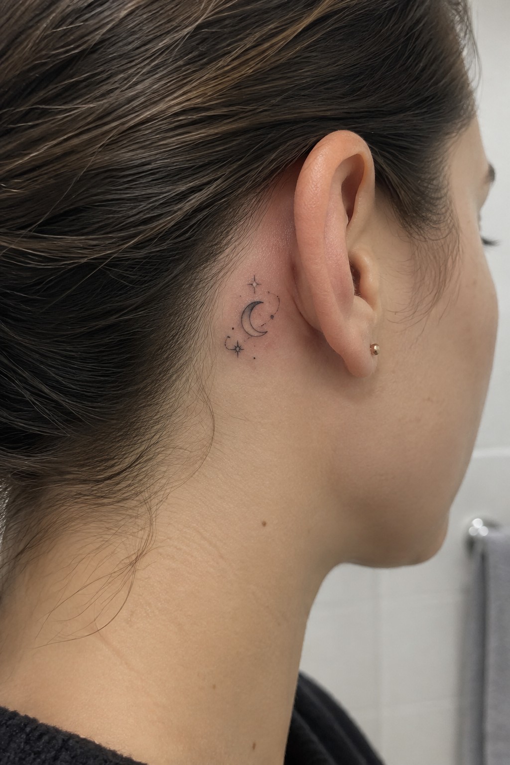 Woman's neck and ear with crescent moon stars tattoo and small gold stud earring.