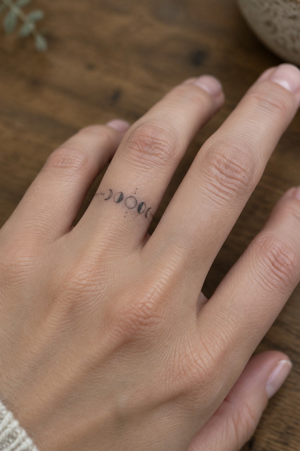 Fine line moon phases tattoo running along the side of a ring finger on a wooden surface.
