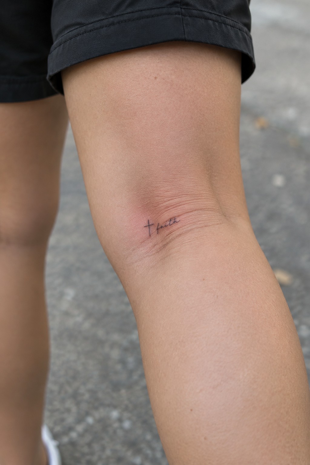Black shorts and small cross faith tattoo on reddened knee over gray pavement