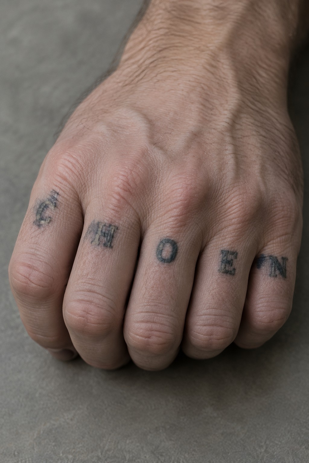Close up of a man's hand showing the word chosen tattooed in black lettering across the fingers.