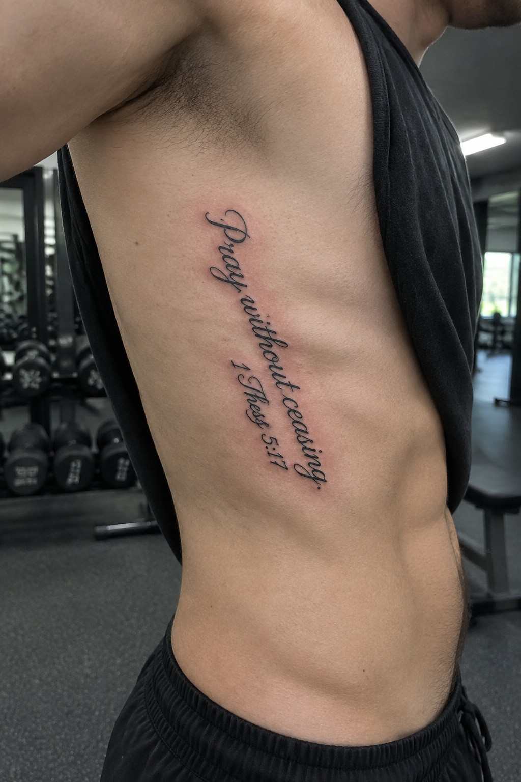 Black cursive tattoo of the bible verse pray without ceasing from 1 thessalonians 5:17 placed vertically on a man's side torso in a gym setting