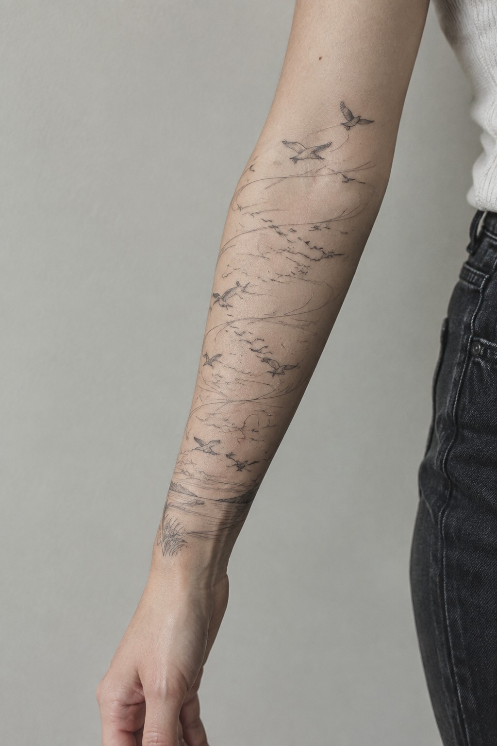 Person's forearm with black line-art tattoo of birds over landscape, white top, dark jeans