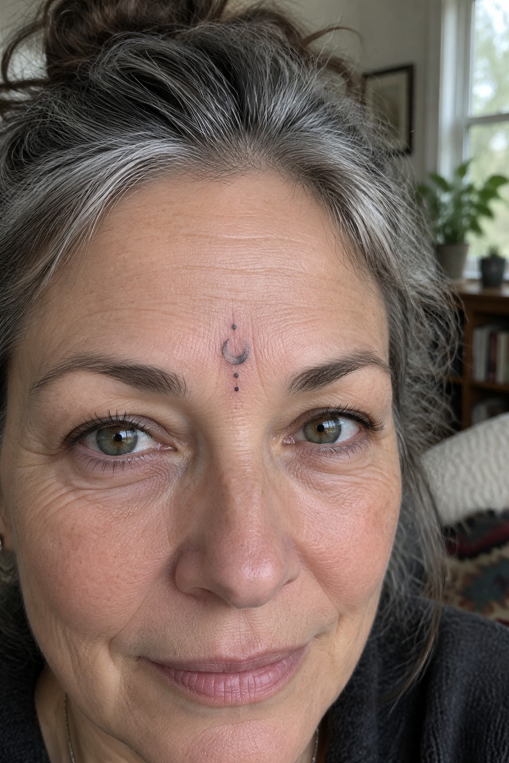 Close up of a small crescent moon tattoo with three dots below it placed between the eyebrows on a woman's forehead