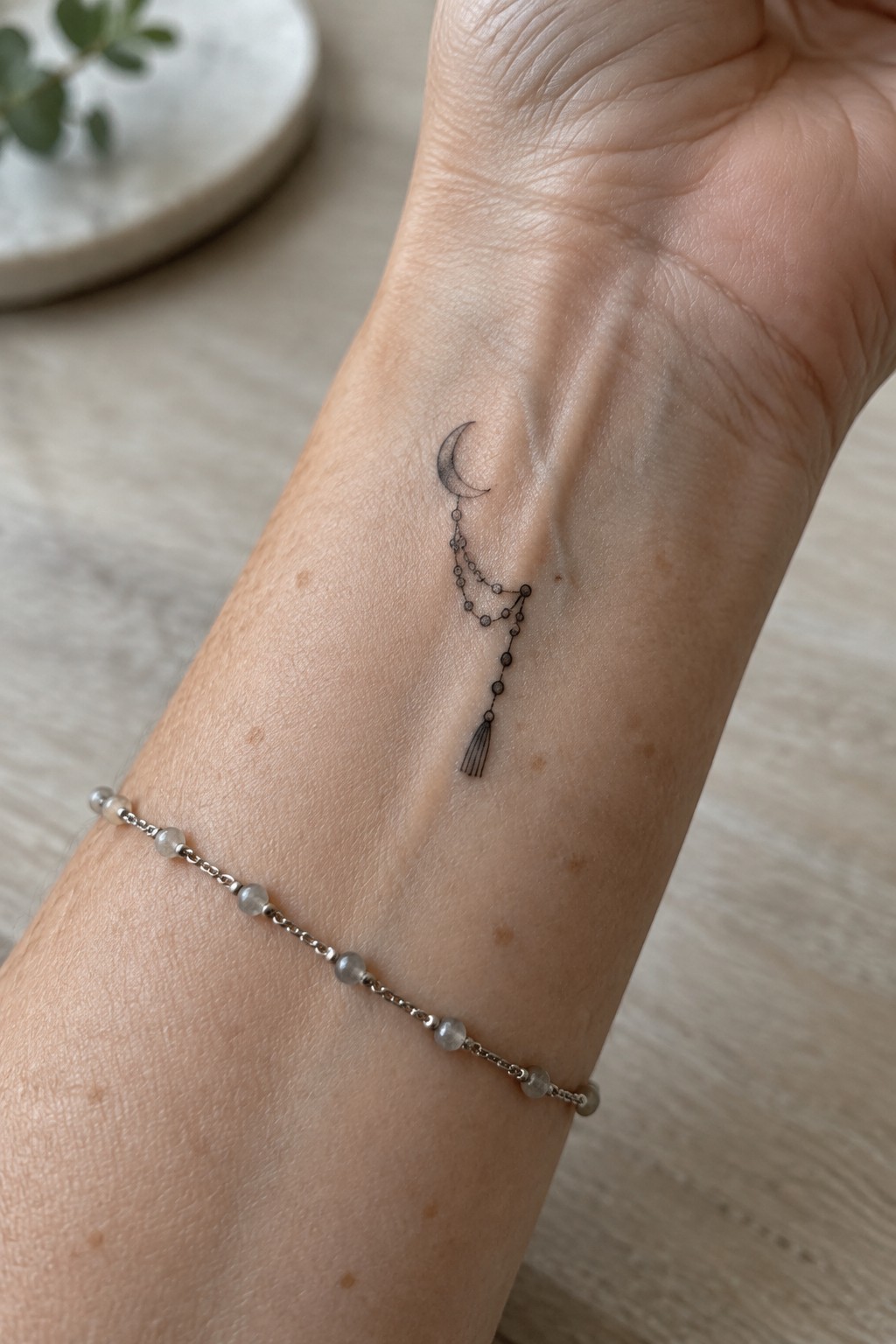 Fine line crescent moon tattoo with dangling beaded chains and tassel on inner wrist next to beaded bracelet