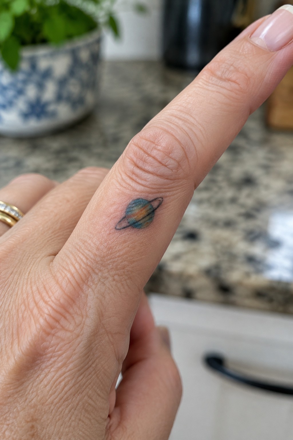 Tiny colorful Saturn tattoo on side of finger with blurred kitchen background