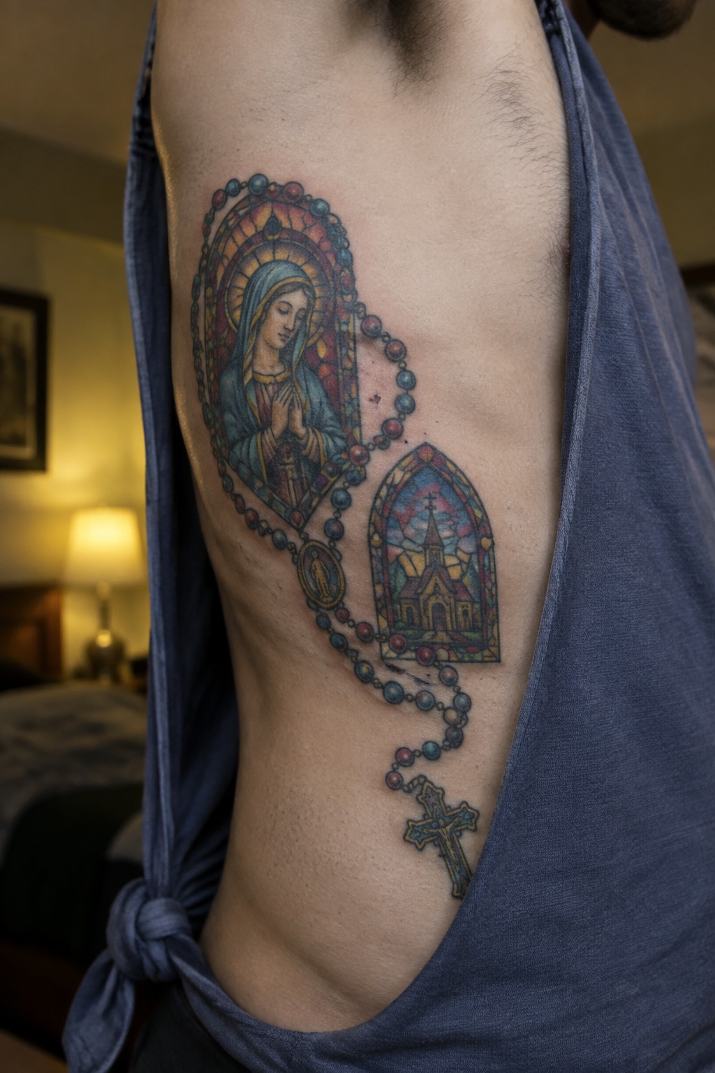Colorful stained-glass Virgin Mary rosary tattoo on torso under blue tied shirt