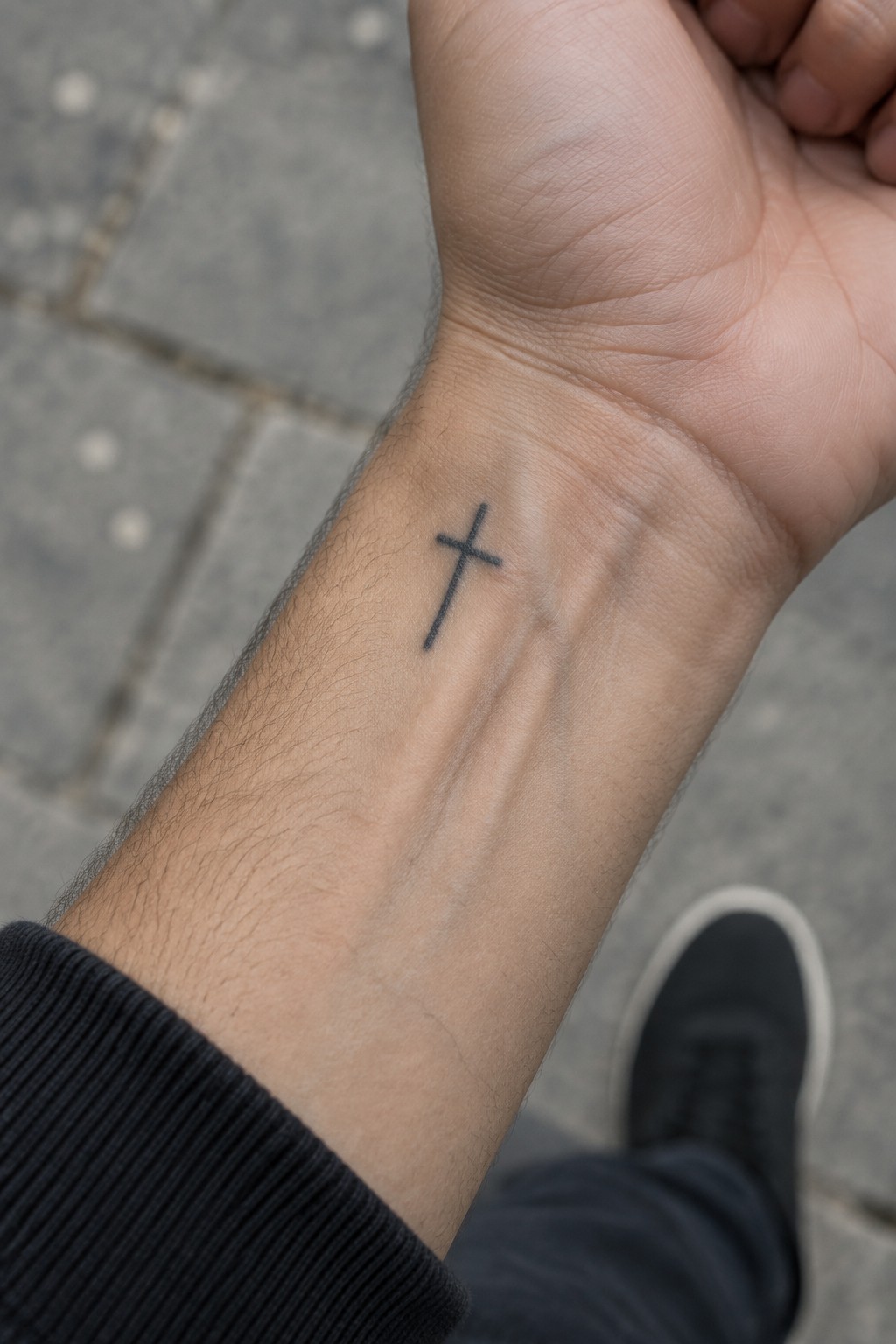 Small black cross tattoo on wrist above black sleeve and shoe on pavement