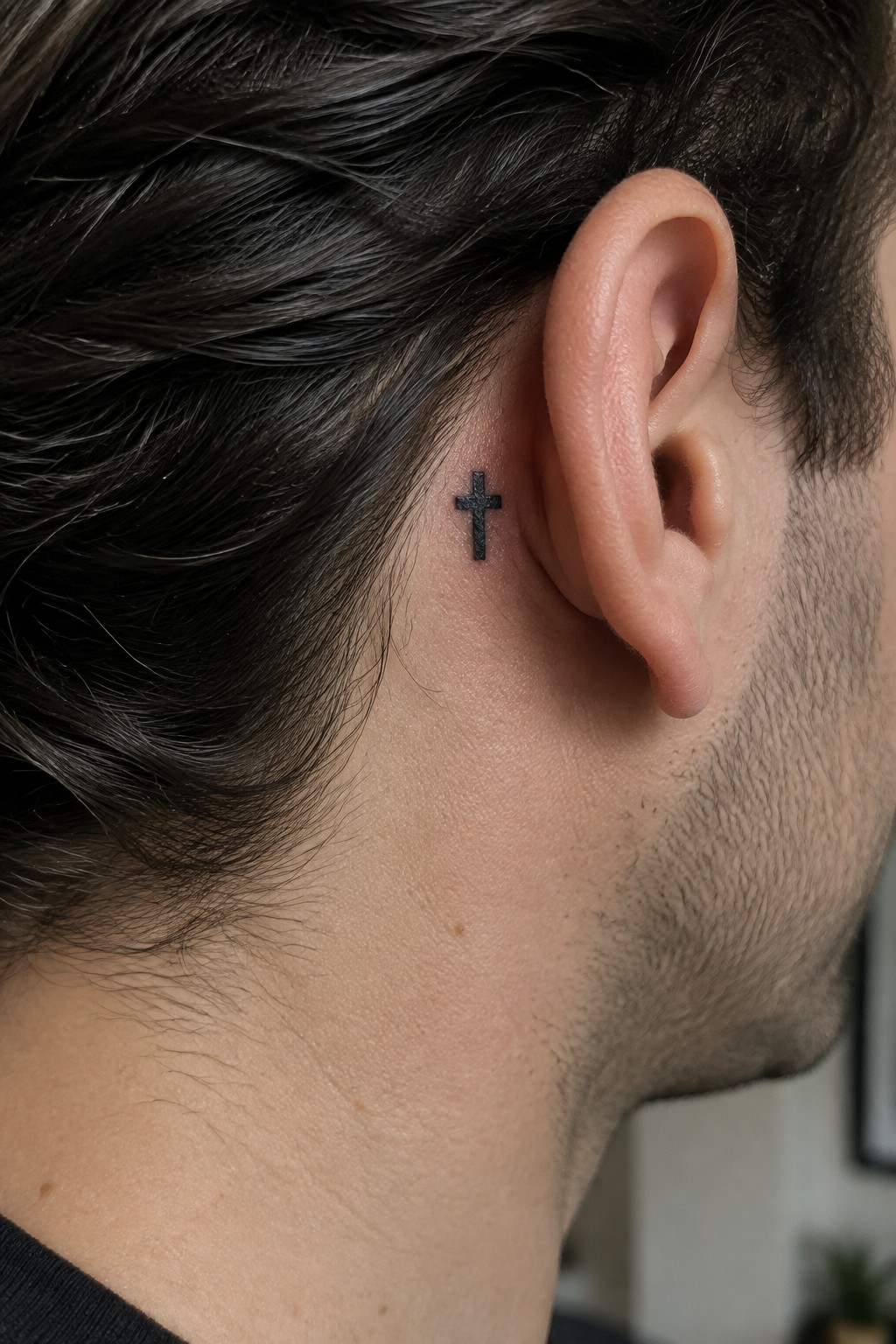 Small black cross tattoo behind the ear on light skin with dark hair.