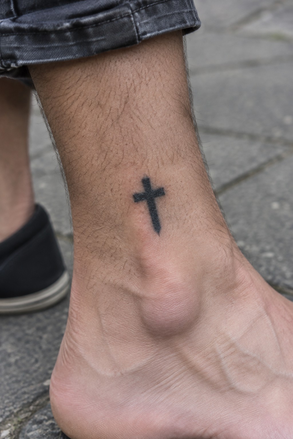 Hairy ankle with small black cross tattoo under rolled dark denim on pavement