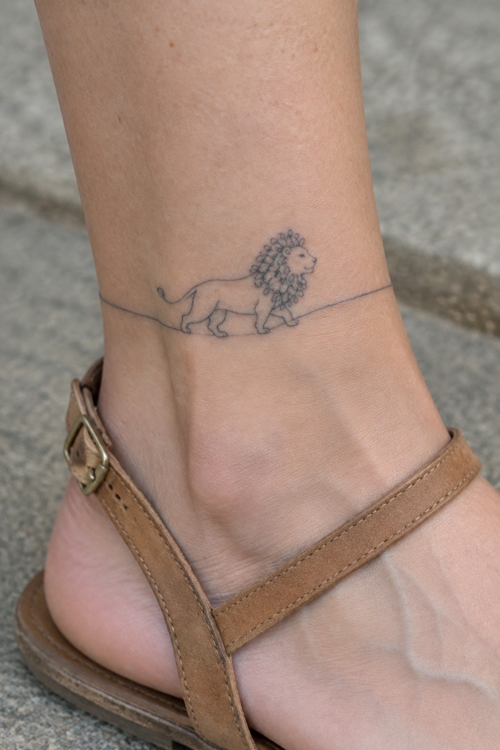 Fine line lion tattoo on the ankle with a thin horizontal line running beneath it, shown above a brown sandal strap.