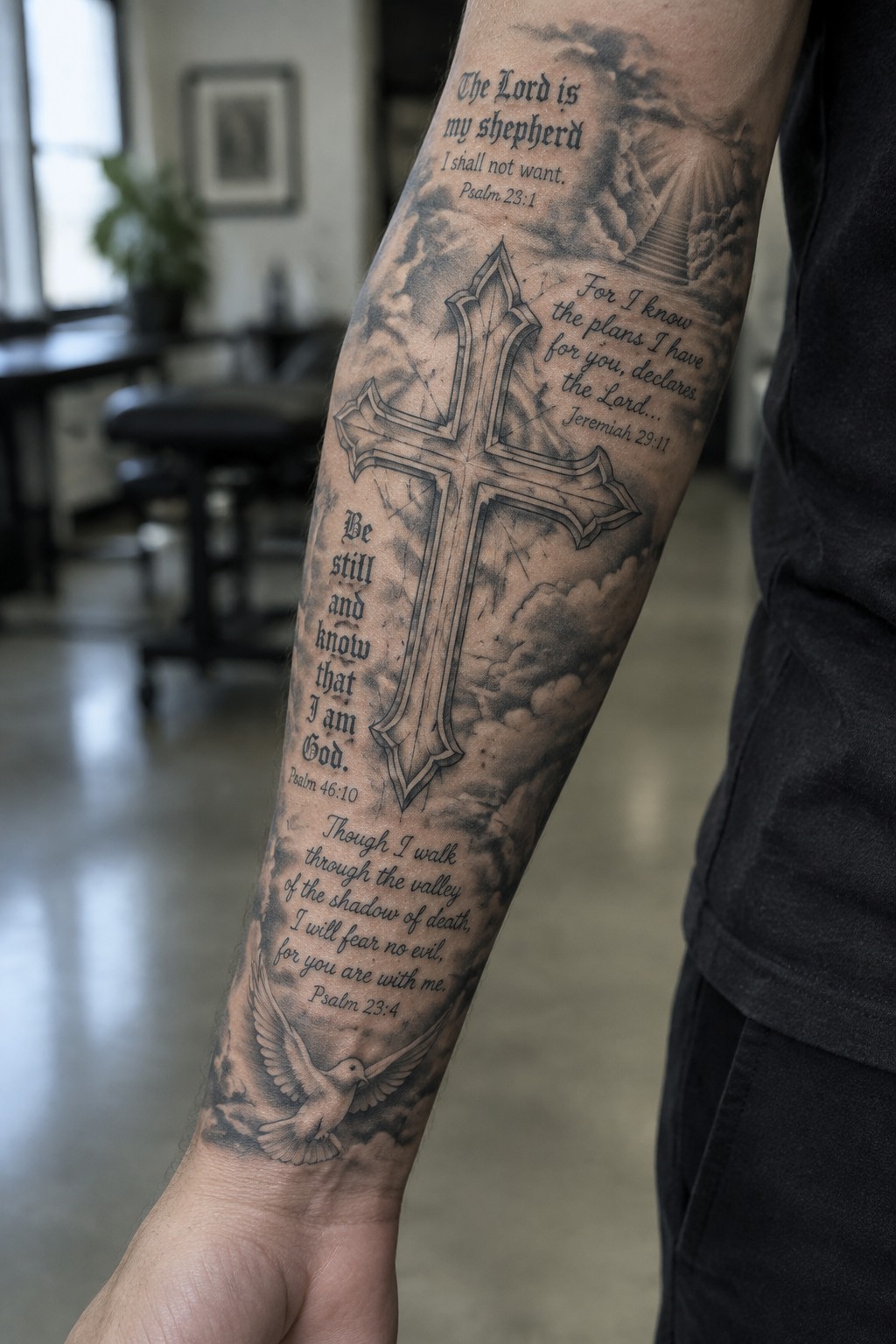 Forearm tattoo showing a large cross with multiple Bible verses and cloud details in black and grey style