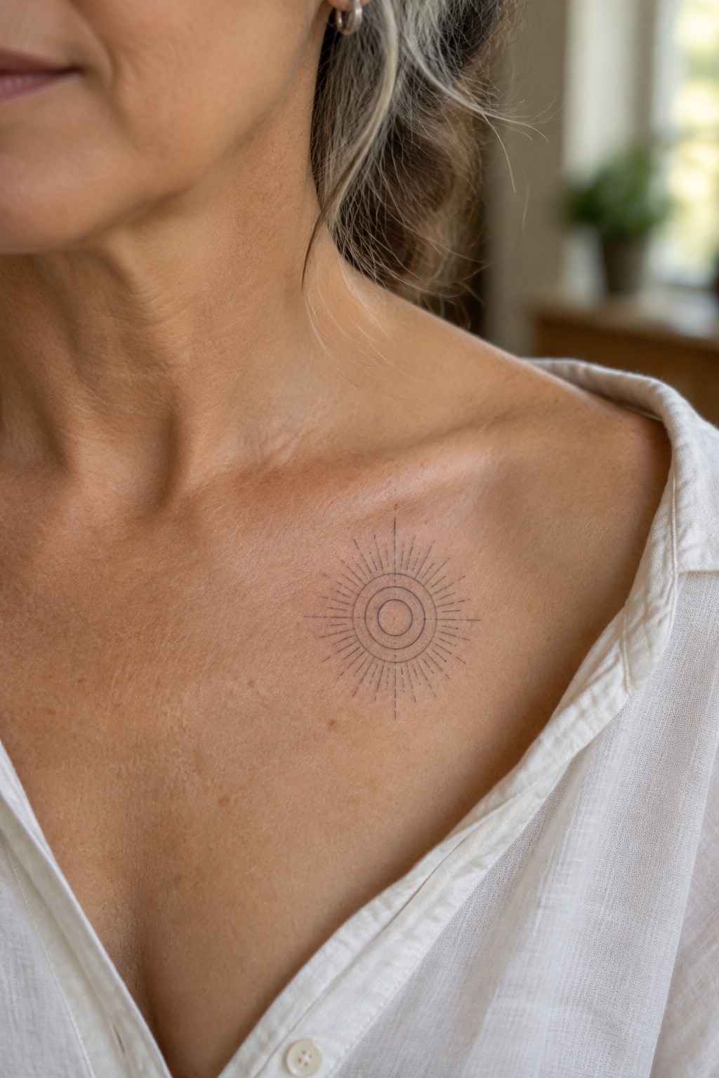 Sunburst tattoo on woman's collarbone above open white linen shirt.