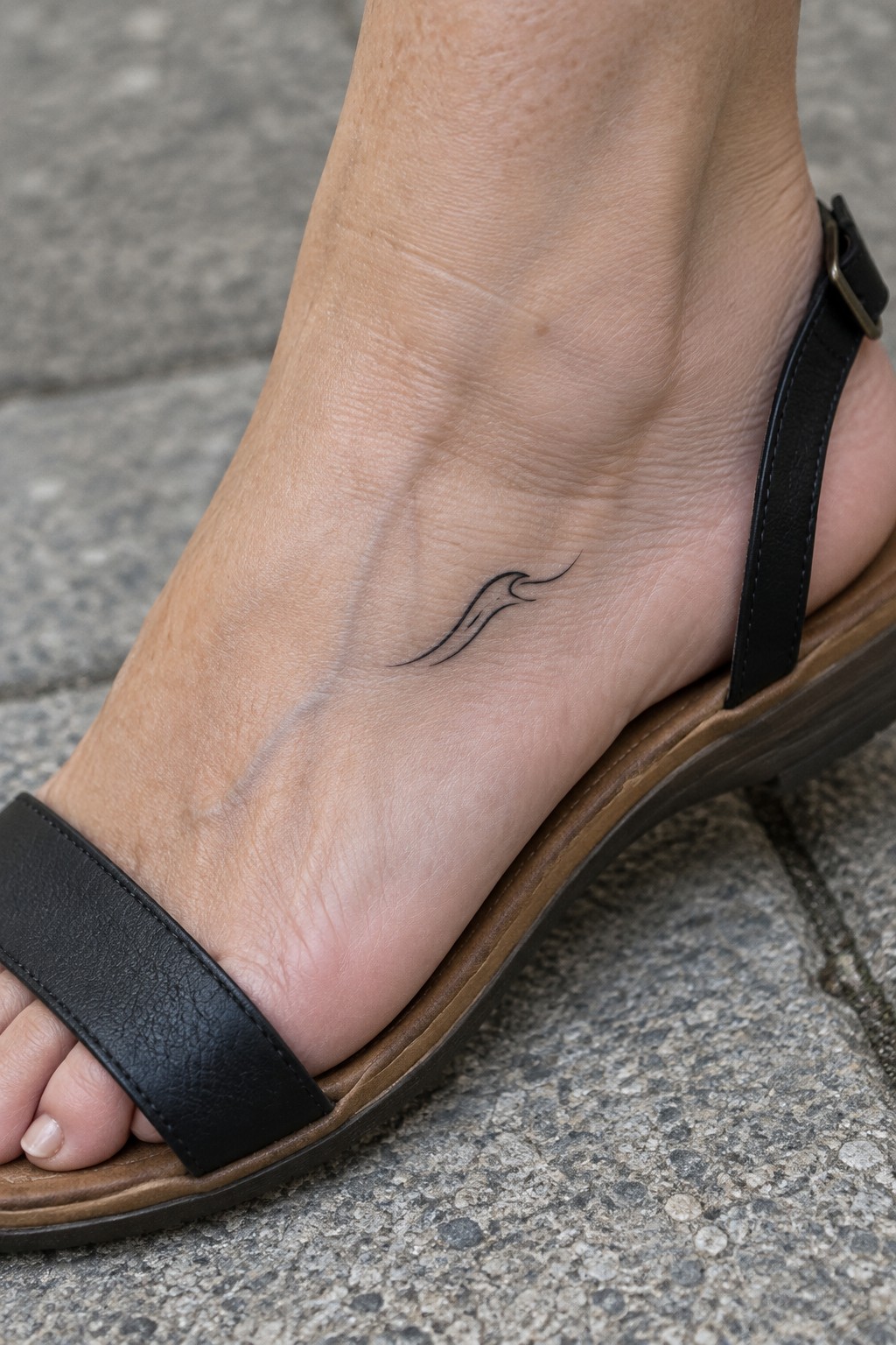 Small fine line flame tattoo on the outer ankle of a foot wearing a black sandal.