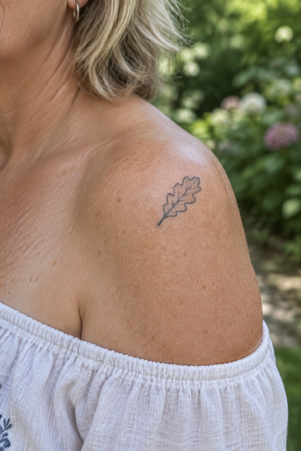 Woman's shoulder with oak leaf tattoo wearing white off-shoulder top in garden