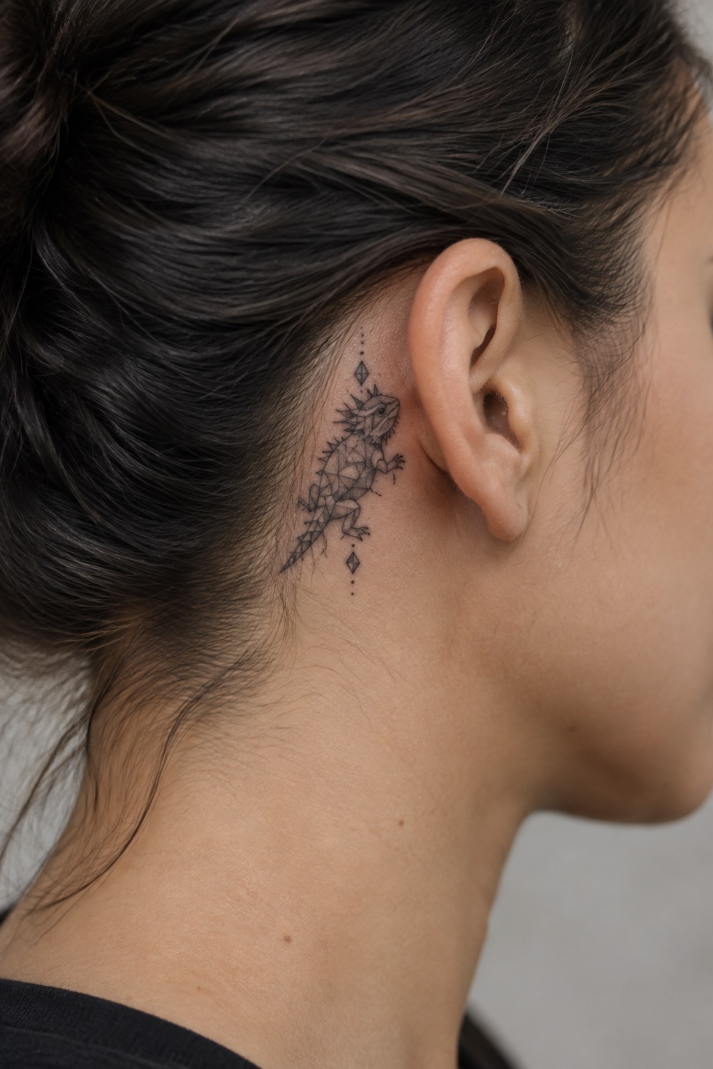 Small geometric lizard tattoo behind a woman's ear on the neck with diamond and dot accents in black ink