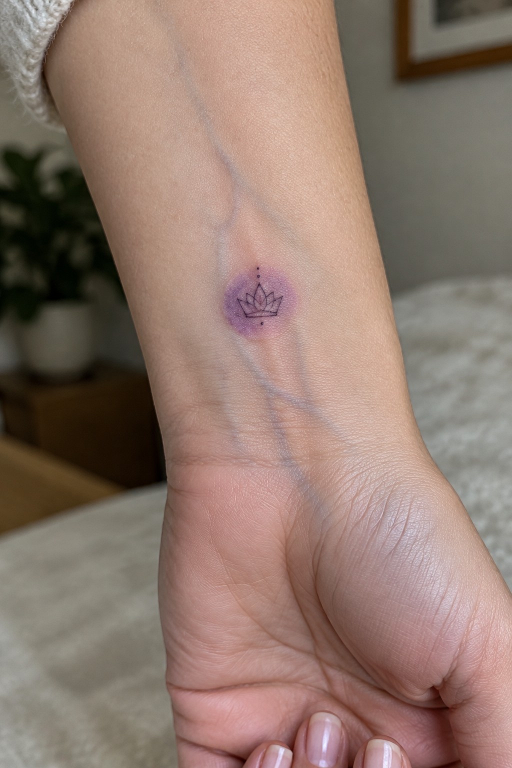 Small lotus crown tattoo with purple watercolor shading on the inner wrist