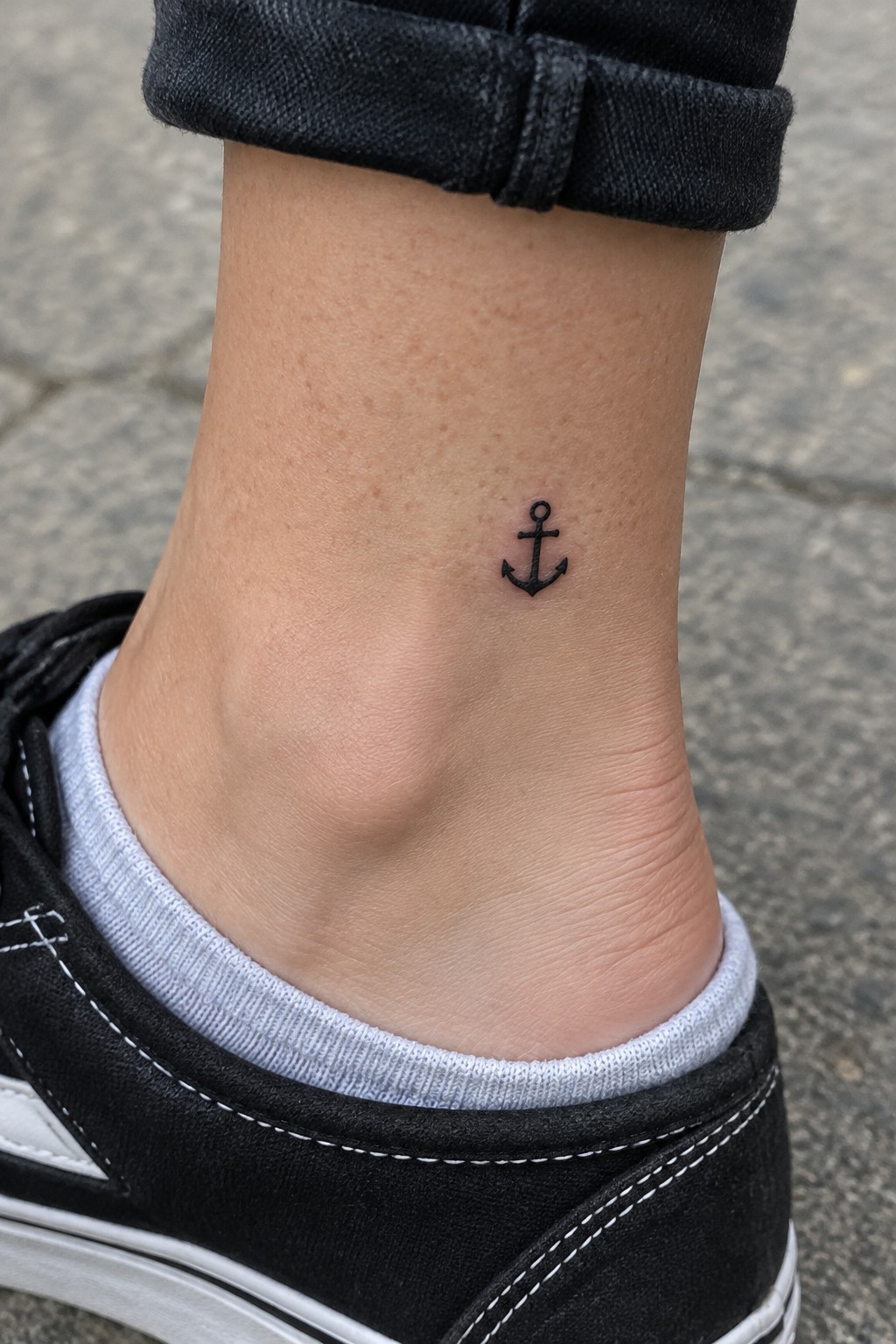 Small black anchor tattoo on ankle above gray sock and cuffed black jeans