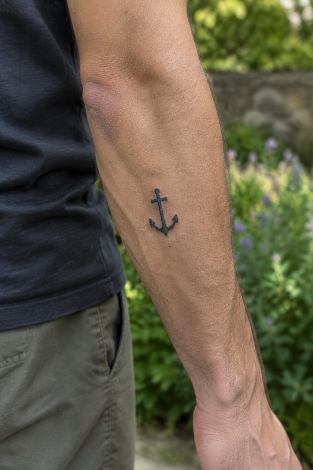 Small black anchor tattoo on the inner forearm of a man wearing a dark t-shirt and khaki pants.