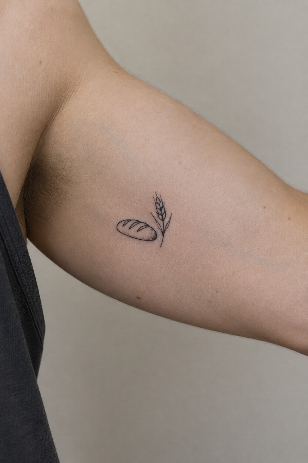 Minimalist black ink tattoo of bread loaf and wheat stalk on upper arm near dark shirt