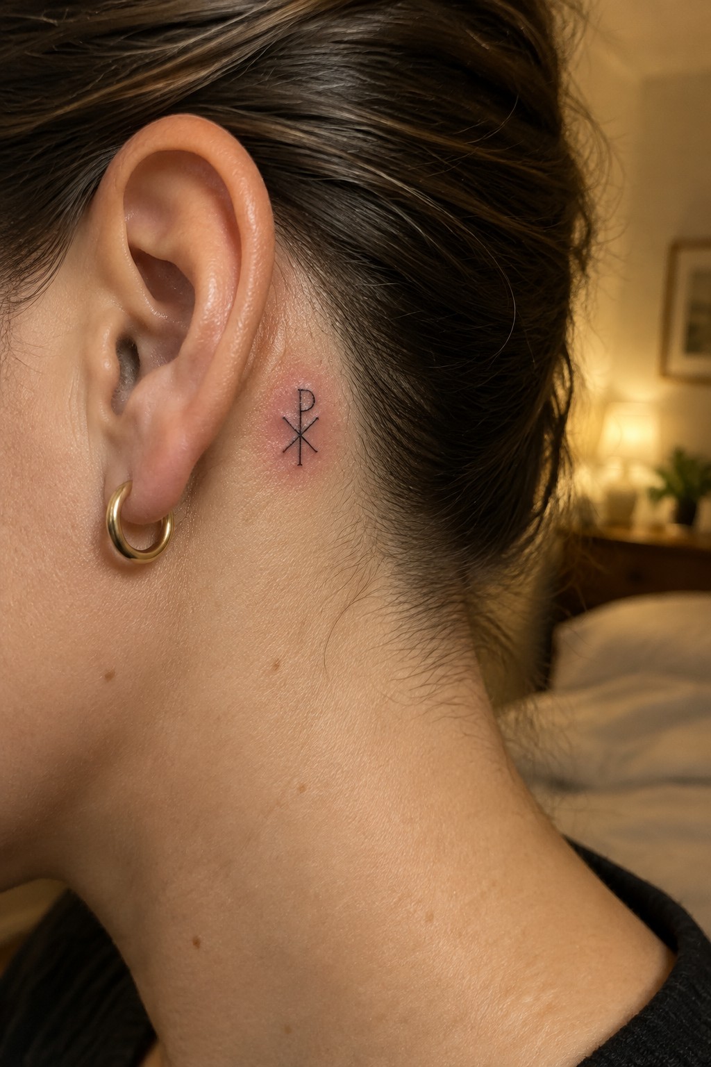 Small black Chi-Rho tattoo behind ear with gold hoop earring
